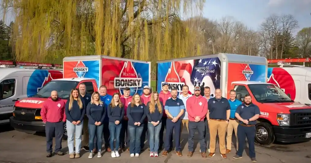 A group of 14 people stands in front of three BONSKY Heating vans on a sunny day, with trees and blue sky in the background.