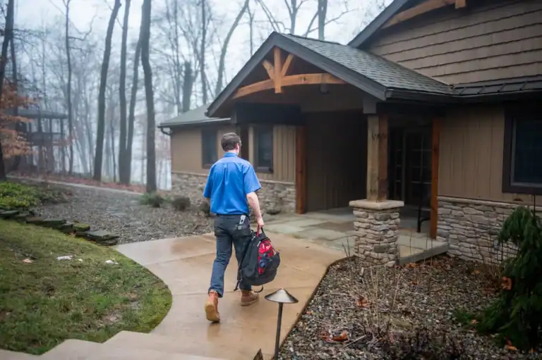 A person in a blue shirt carrying a tool bag walks on a curved path toward the front door of a house surrounded by trees on a foggy day.